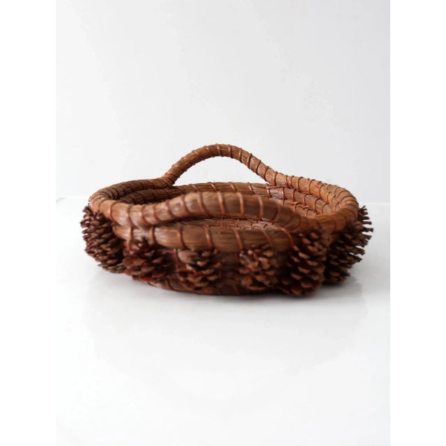 Crafted from coiled pine needles and trimmed with small pine cones, this vintage folk art basket has an earthy, organic...