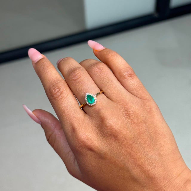 This halo ring showcases 0.7 carats of natural pear-cut emerald, set in 18K yellow gold. The center stone measuring 7 x 5...