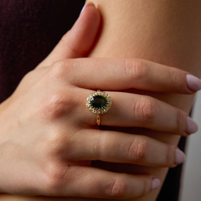 A captivating Victorian cluster ring centered around a richly colored oval green sapphire weighing 2.10 carats. The...