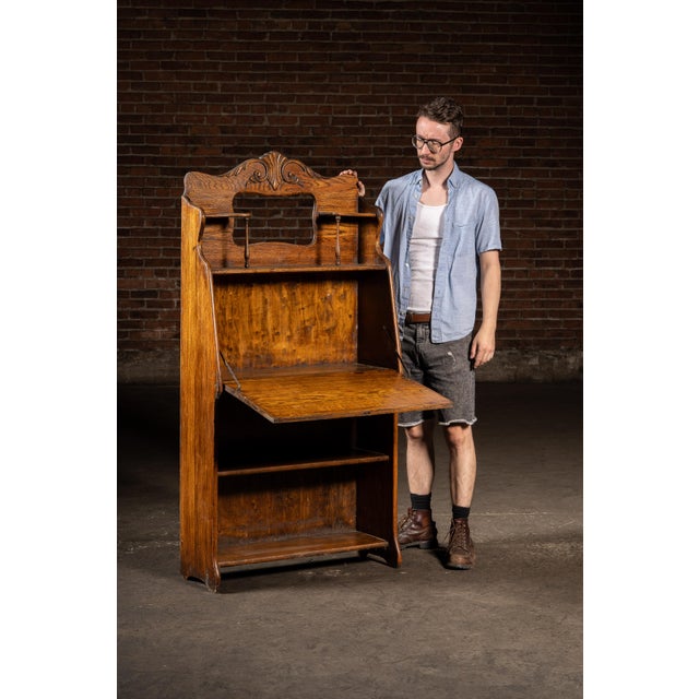 Solid oak drop-front secretary featuring a carved foliate crest, turned spindle supports, and open shelving beneath the...