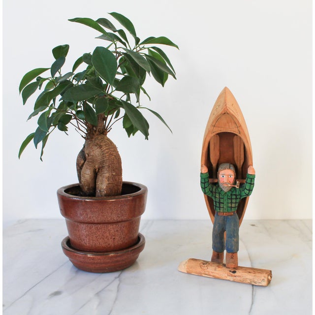 Vintage hand-carved wooden figurine of a man in a plaid shirt, smoking a pipe and carrying a canoe. Some minor age wear.