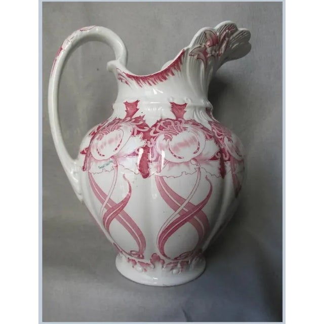 An Antique (circa 1910-1919) Art Nouveau Water Pitcher. The Water Pitcher is made of pottery and features a red transfer...