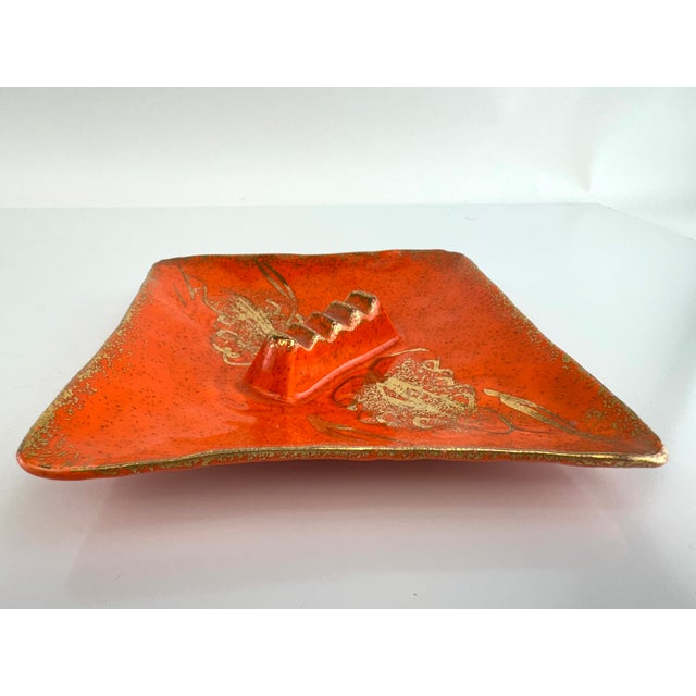 A mid-century ceramic ashtray featuring a vibrant red-orange glaze accented with gilt floral motifs and subtle speckling...