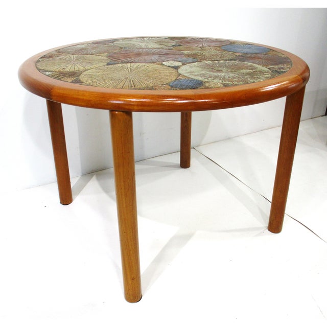 A darker teak wood framed round dining table with pole styled legs and having a colorful earth tones and blue detailed...
