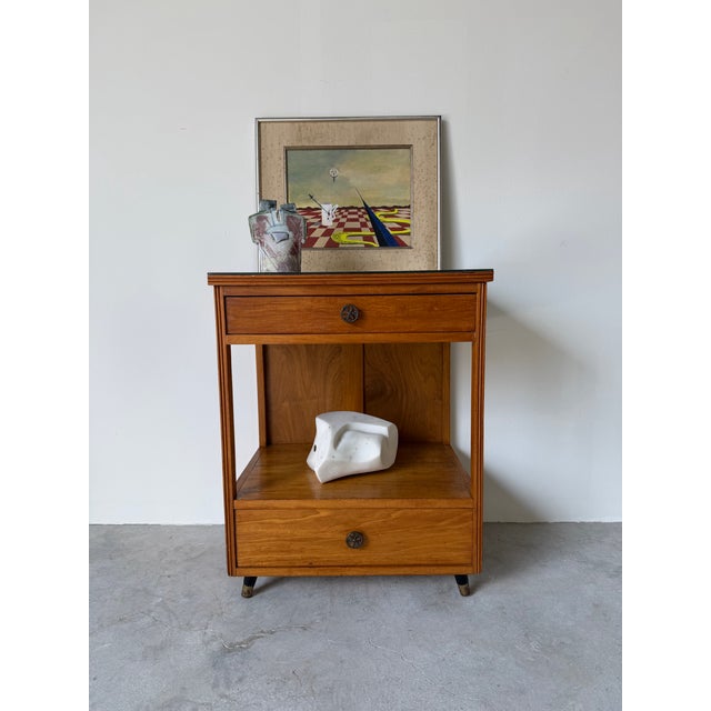 A clean and functional 1960s Mid-Century Modern nightstand featuring warm wood tones and a minimalist open-frame design....