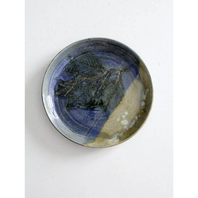 This is a signed studio pottery plate. The hand-crafted plate features a leaf and branch imprint with a striking blue and...