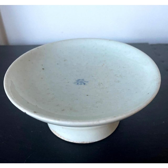 A small ceramic stemmed dish with high cylindrical foot in white glaze with a cobalt blue underglaze inscription in the...