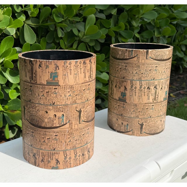Pair of vintage cylindrical wastebaskets featuring detailed Egyptian Revival imagery with hieroglyphic motifs, figures,...
