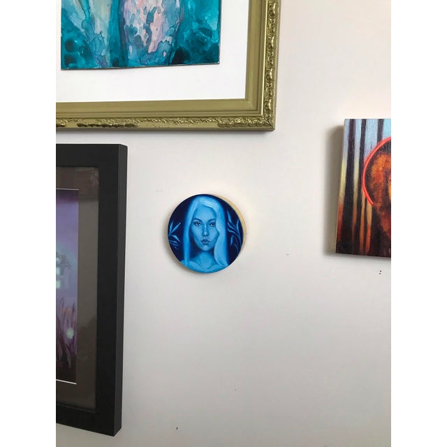 This circular wood tondo portrays a striking figure with ethereal blue tones, capturing a serene yet enigmatic expression....