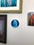 This circular wood tondo portrays a striking figure with ethereal blue tones, capturing a serene yet enigmatic expression....