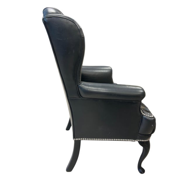 This is a classic, high-back wingback armchair upholstered in black leather. The backrest features deep button tufting in...