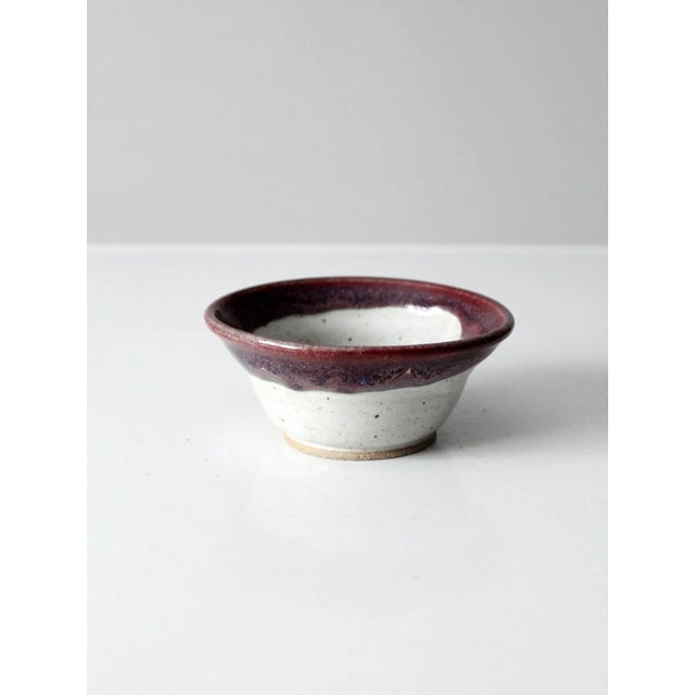 This is a vintage studio pottery bowl. The hand-crafted bowl features a white speckled glaze with a rich burgundy tone...