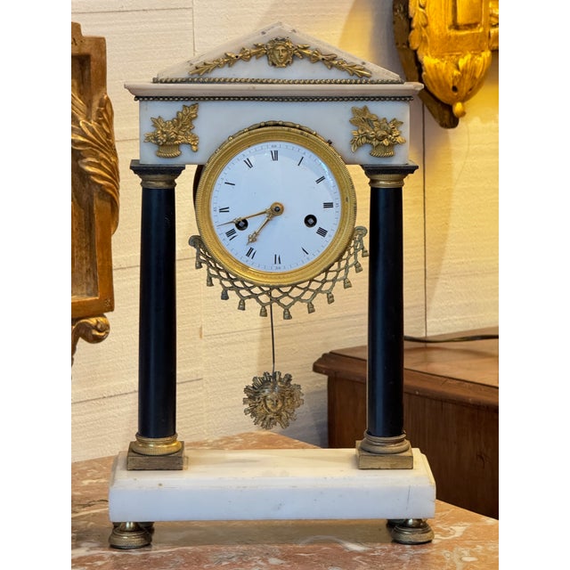 French mantel clock in the Empire style, featuring a white marble case with black column supports and classical gilt...