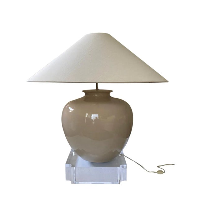 A large beige ceramic urn with an oversized linen coolie shade sits on a dramatic 4” thick Lucite base with curved notched...