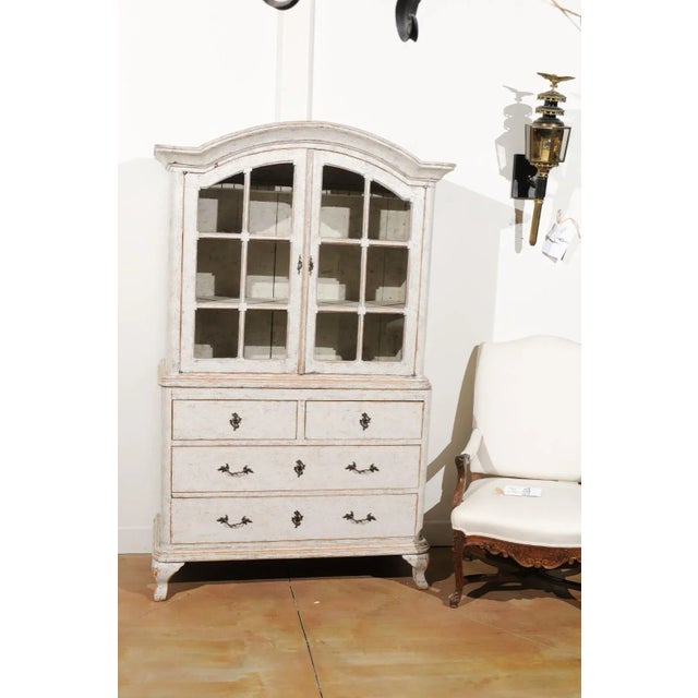 A Swedish Rococo style painted wood vitrine from the 19th century, with bonnet top and glass doors. Born in Sweden during...