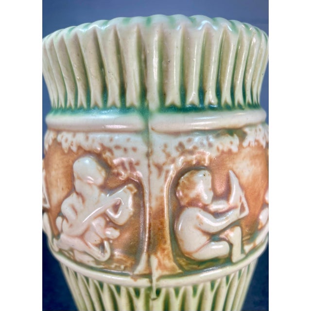 Green art pottery vase featuring trees, ivory flutes, & cherubs on brown band. C. 1915.