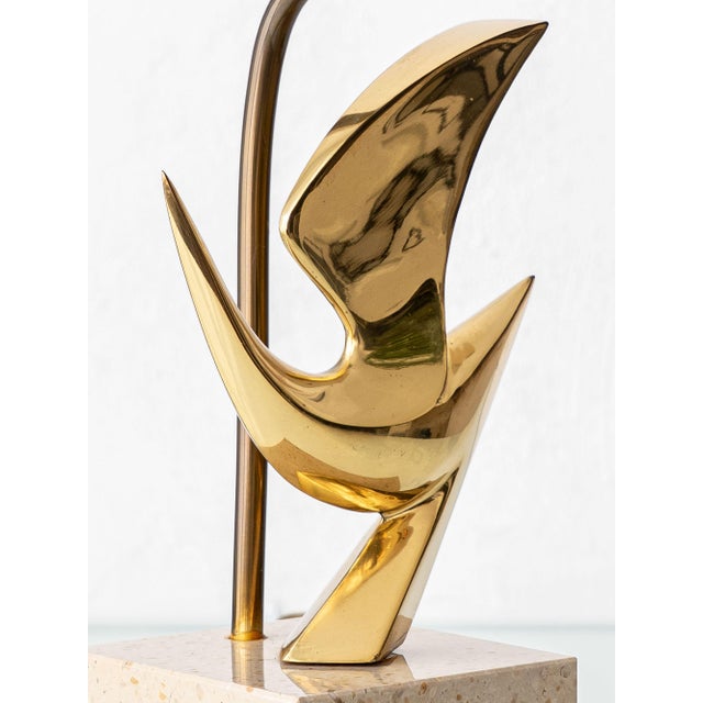 A sculptural 1980 table lamp by Philippe Jean, featuring a polished brass abstract bird mounted on a travertine pedestal...