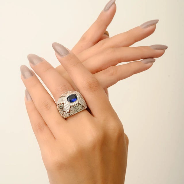 This bold 925 sterling silver ring features an oval-cut blue stone at the center, framed by round-cut white cubic zirconia...