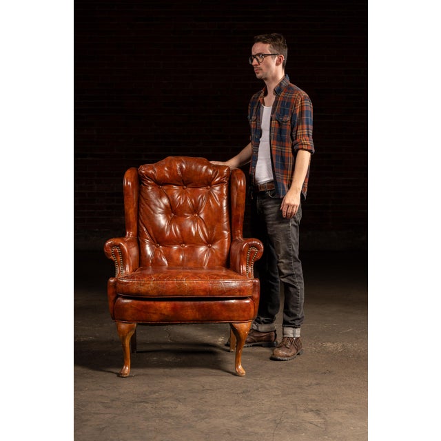 Tufted leather wingback armchair in a rich burnt amber hue, featuring brass nailhead trim and cabriole front legs. The...
