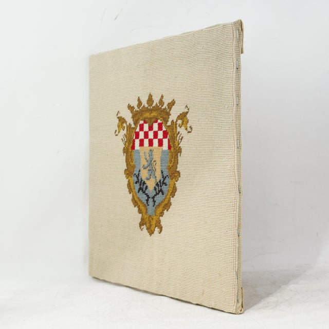 This elegant vintage needlework piece features a meticulously heraldic crest centered on a neutral cream textile...