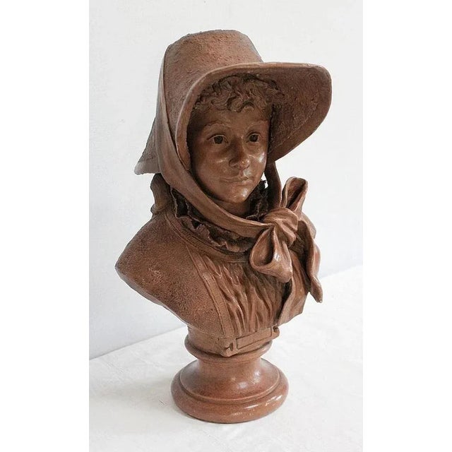 Terracotta bust representing a woman with a youthful face and fine features. She is wearing a generous hat, with soft...