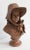 Terracotta bust representing a woman with a youthful face and fine features. She is wearing a generous hat, with soft...