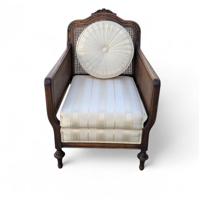 French provincial style cane armchair with a carved wood frame, woven cane panels, cream striped upholstery, and round...