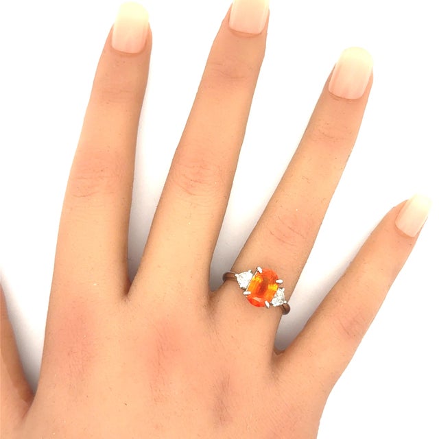 A striking three-stone ring featuring an exquisite oval-shaped orange sapphire, weighing 3.89 carats with one of the...