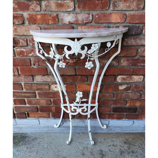 A charming petite demi-lune console table featuring an ornate white metal base with delicate floral and scroll motifs and...