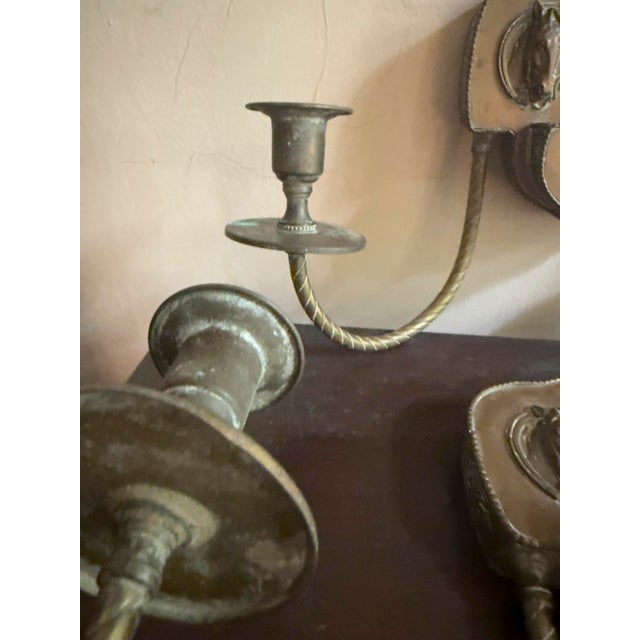 Pair of 1970s vintage Colonial Revival brass wall sconces with double candle arms and horse head relief mounts. Each...