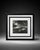 Elegant black and white framed landscape photograph featuring a small boat, tree, shoreline, and dramatic outdoor scenery....