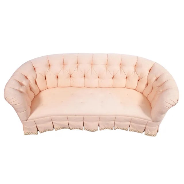 A vintage pale pink button tufted upholstered sofa with a pleated skirt. The sofa back is completely tufted and curves...