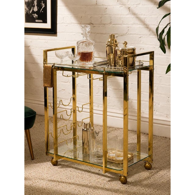 A glamorous vintage bar cart featuring a polished brass-tone metal frame with clean architectural lines and an open, airy...