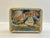 A vintage tin box with a tropical sunset scene with a masted sailing ship in full sail against a backdrop of clouds.