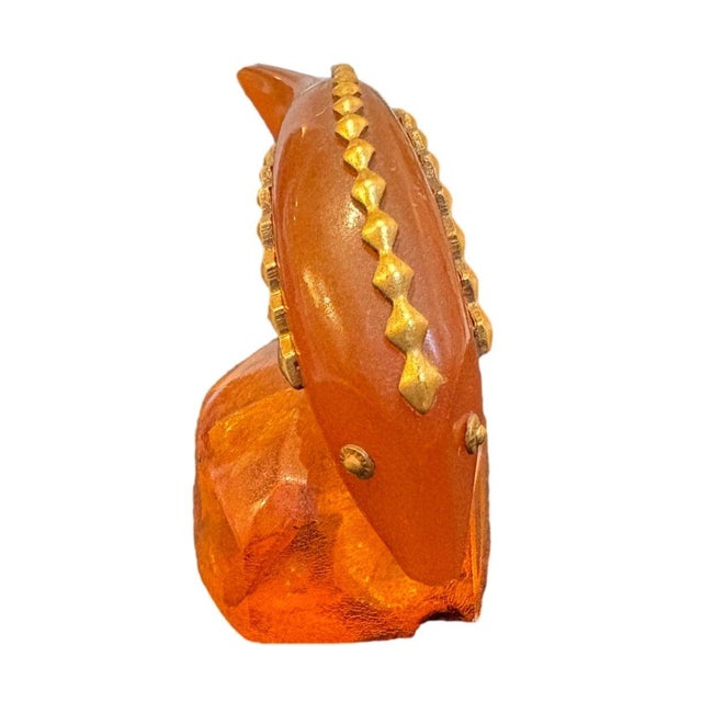 This striking amber figurine features a curved fishtail-like form adorned with intricate brass detailing along its ridges....