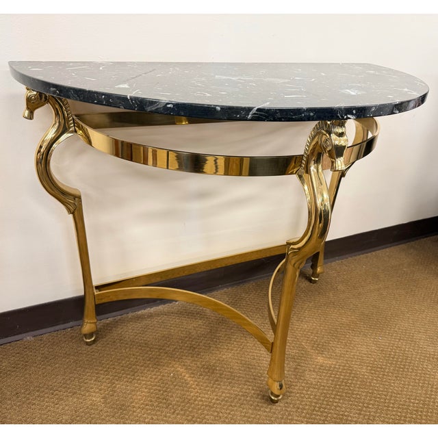 A stylish brass demilune console table with a black marble top and distinctive horse-head supports, offering a...