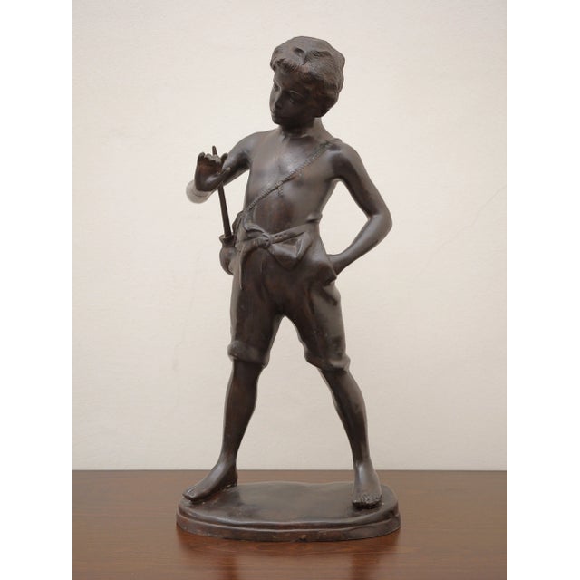 A decorative bronze statue representing a standing boy in a slightly relaxed posture, with his head tilted to his right...