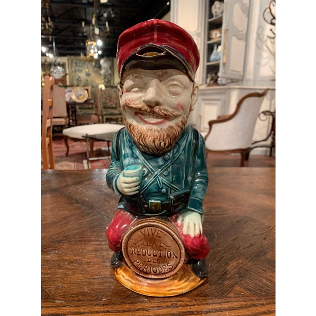 Colorful and highly expressive, this antique barbotine pitcher depicts a Belgian soldier seated confidently upon a barrel....