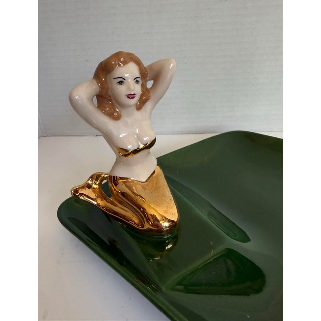 This charming vintage figural ashtray features a glamorous mermaid reclining along the edge of a sculptural emerald green...