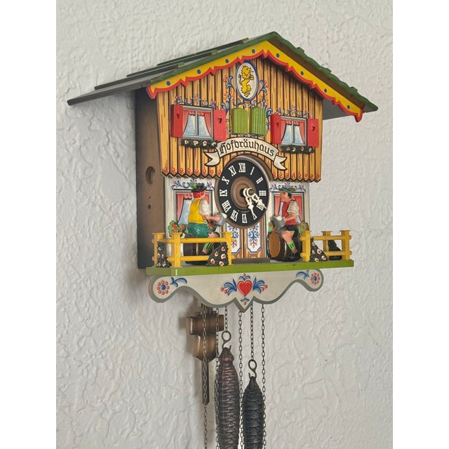 A 1970s West German Hofbräuhaus chalet cuckoo clock with a yellow body, green roof, lion emblem, beer drinkers on the...