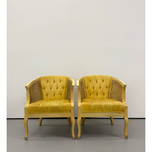 A distinctive pair of vintage barrel-back armchairs featuring warm cream-painted wood frames, woven cane side panels, and...