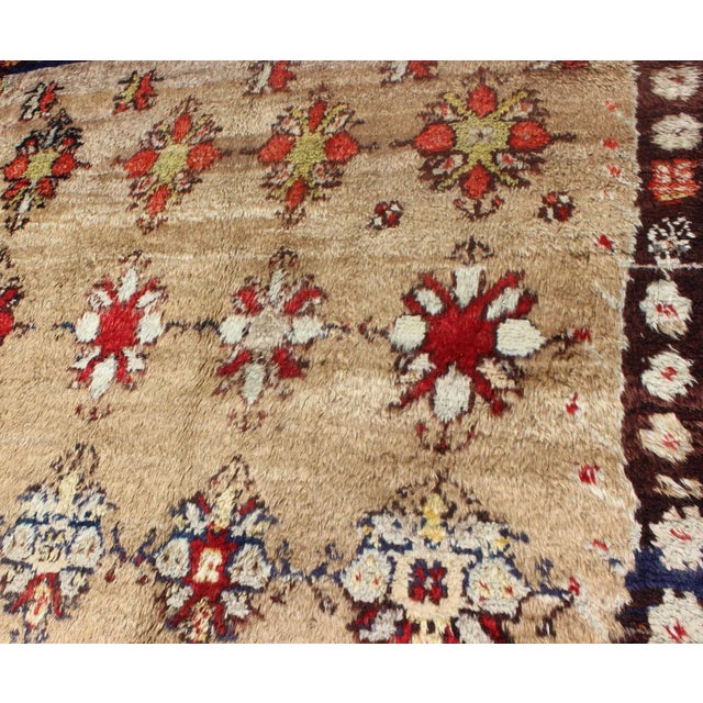 This Tulu carpet features several multicolored floral motifs laid across a sand-colored field and enclosed within a...