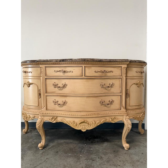 A stunning vintage French Provincial–style marble-top credenza featuring an elegant curved silhouette, ornate carved...