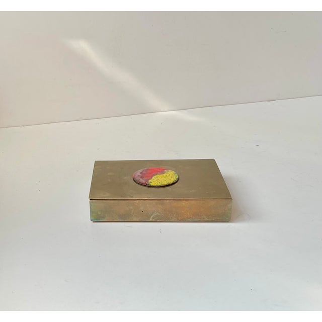 A brass cigarette or cigar box executed entirely of brass and set with an unusual and vivid circular enamel plaque in red...