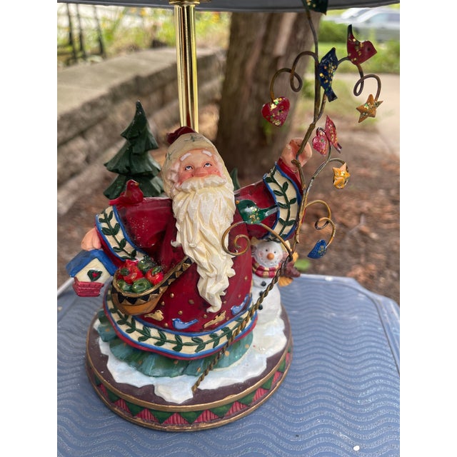 Whimsical Christmas table lamp features a hand-painted resin base depicting Santa Claus carrying a birdhouse and basket of...