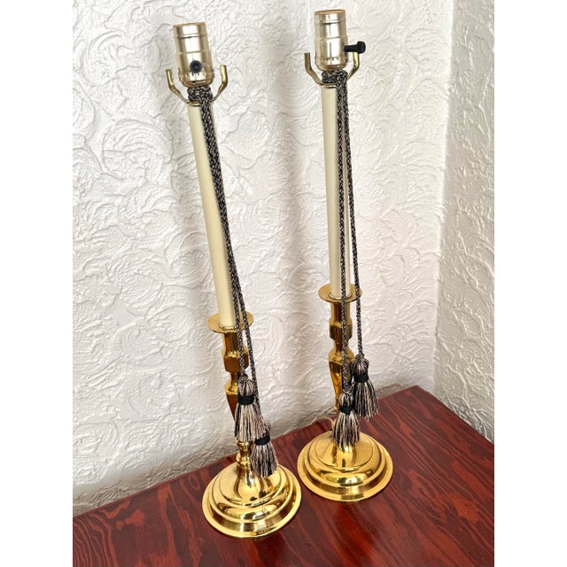Pair of brass candlestick table lamps with braided tassel cords. Each lamp has a tall cream candle sleeve on a slender...