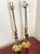 Pair of brass candlestick table lamps with braided tassel cords. Each lamp has a tall cream candle sleeve on a slender...