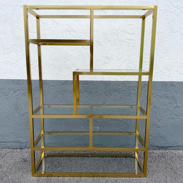 This striking Hollywood Regency étagère features a clean geometric brass-tone frame paired with clear glass shelves,...