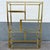 This striking Hollywood Regency étagère features a clean geometric brass-tone frame paired with clear glass shelves,...