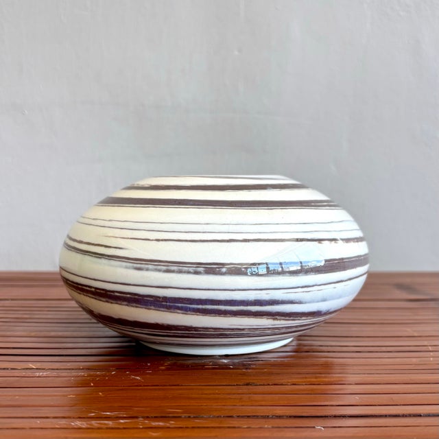 This low, rounded vessel features a creamy glaze accented by hand-drawn brown rings that spiral organically around its...